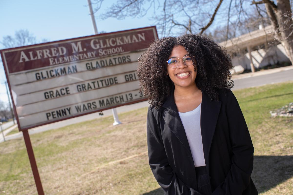 Reach to Teach alumna Lisha standing outside school smiling wearing a black coat, white shirt, and glasses.