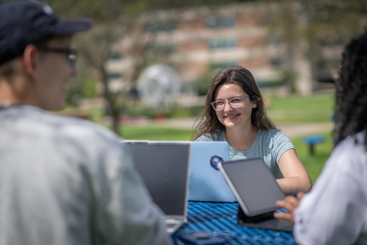 Three students sitting outside on a sunny day, working on laptops.