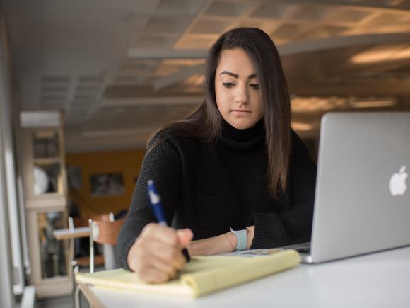 Graduate student writing with laptop on desk.