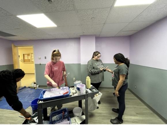 Students creating a pediatric waiting room at MiraVista Behavioral Health Center in Holyoke, MA, as part of the nursing department's the Psychiatric Nursing class. Four students are painting a white room to green, and use paints provided by a small, portable, black worktable.