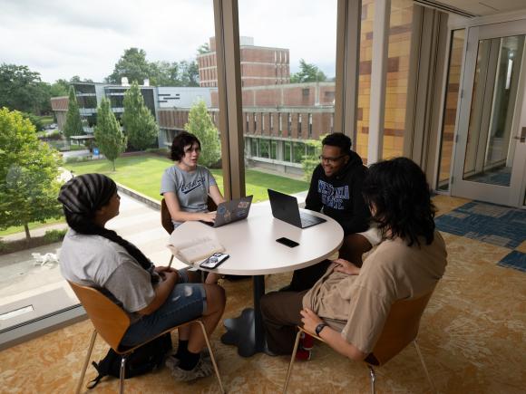 Group of students sitting around a round table chatting with laptops.