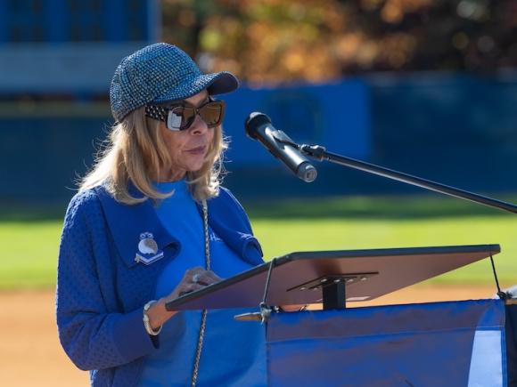 President Linda Thompson of Westfield State giving an address for this year's Homecoming. She's in all blue and wears a blue cap while speaking at a podium on an athletic field.