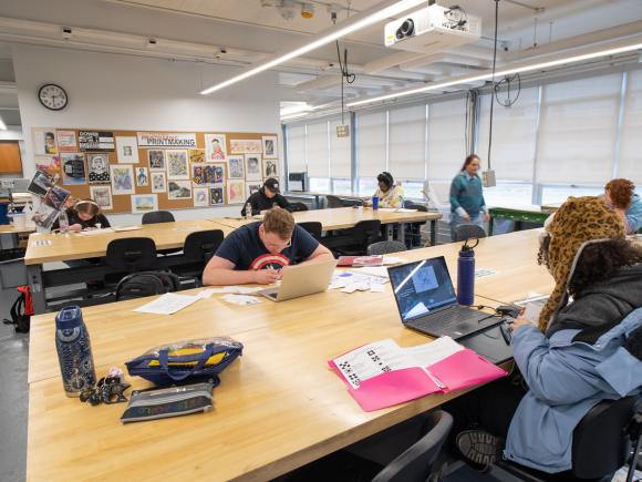 Students in an art class working on projects with laptops on tables.