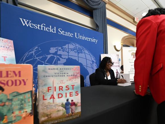 Woman signs books at a table while speaking to another woman