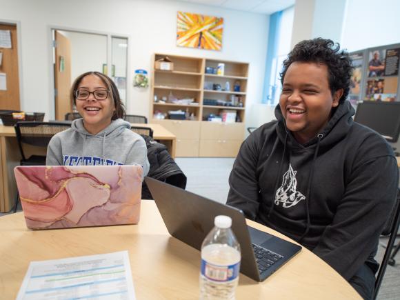 Two computer science students sitting side by side with laptops open, laughing.