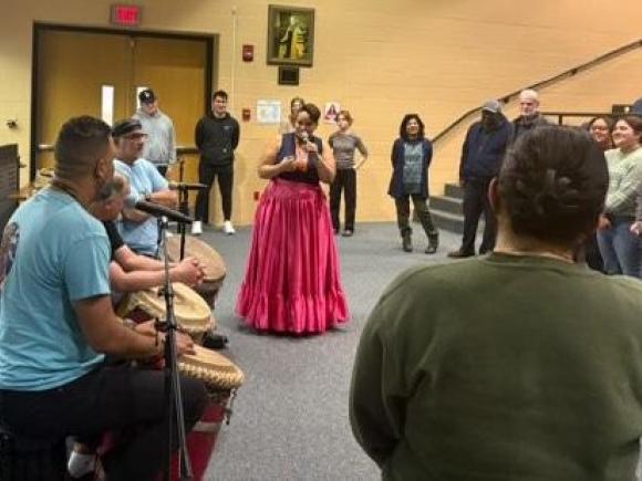 Woman sings to audience with row of drummers and dancers