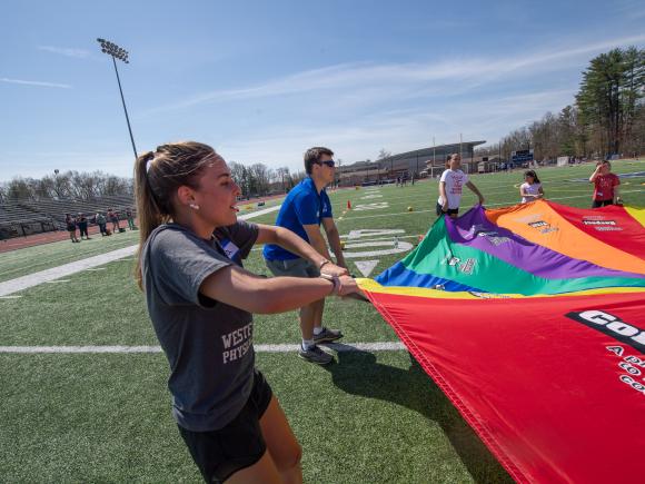 A college student helping children play with a colorful parachute during a Physical Education Day event.