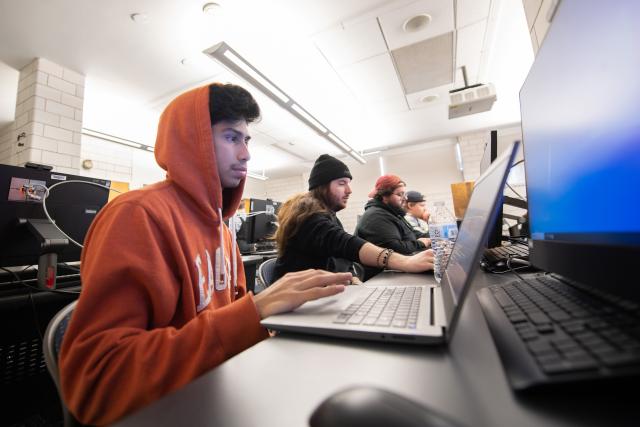 CIS student wearing orange hoodie working on laptop in classroom.