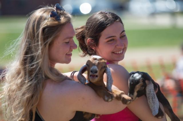 Fresh Check Day 2023. Two women students hold baby goats as part of learning about self-care. One has long, blond hair while the other has brown hair in a ponytail. The goats have brown fur and are nestled in their arms.