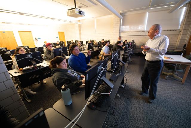 Students listening to a professor speak in front of a classroom.