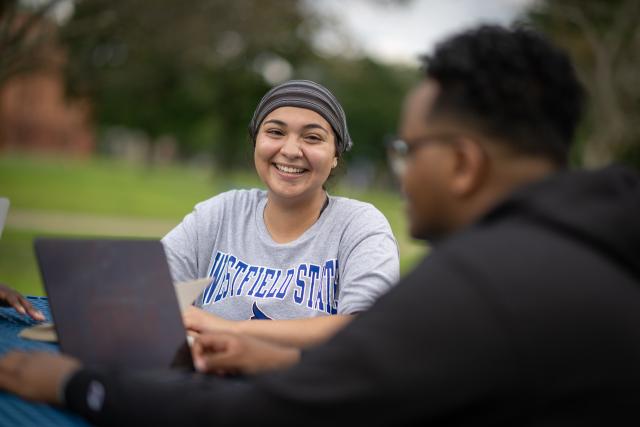 Student smiling outside on campus green with laptop.