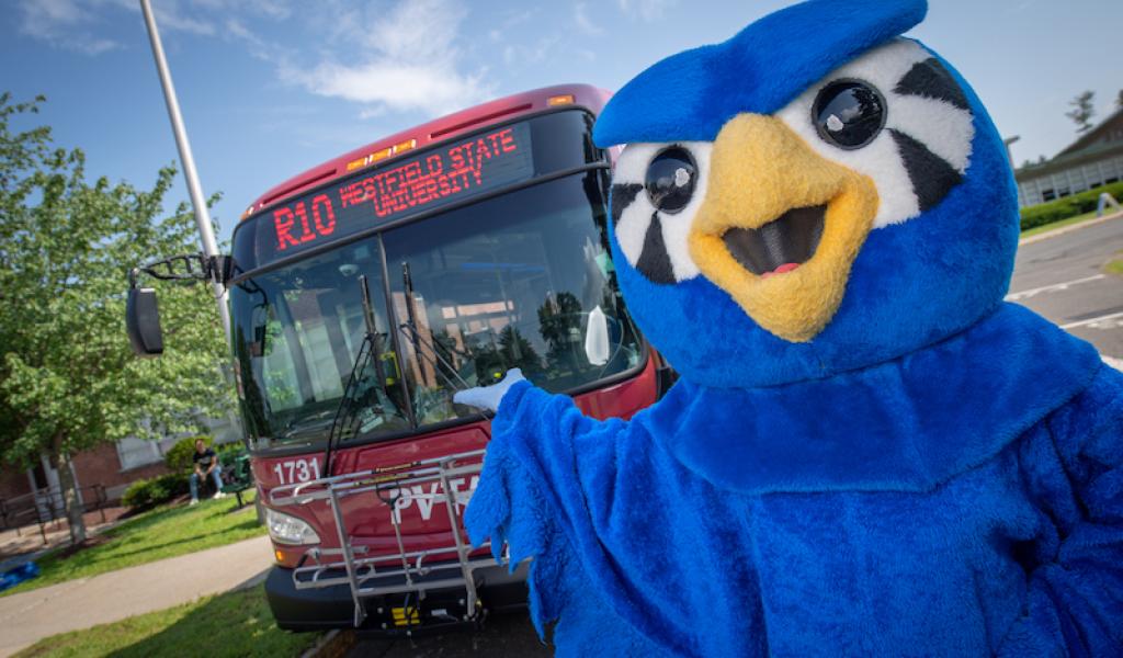 A photo of Nestor in his blue, owl costume and pointing towards a red bus which has the university's address and route on the front.