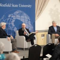 Three men sit in chairs on a stage in front of an audience