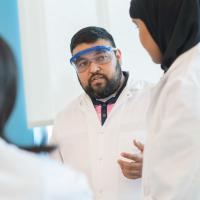 Roderico Acevedo teaches in a lab coat and goggles while two students also in lab coats look on